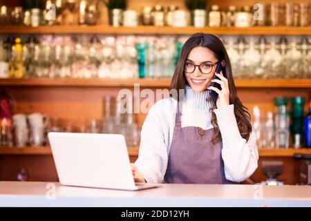 Portrait of a beautiful waitress using mobile phone and laptop while staning behind the counter at a restaurant. Small business. Stock Photo