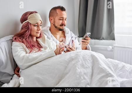 relaxed couple use mobile phone lying on bed together, caucasian man and woman happy to have rest, look at screen of mobile phone Stock Photo