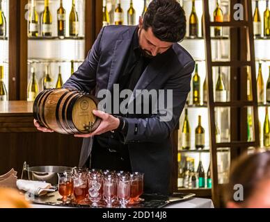 A bartender at the Hotel Alàbriga near Sant Feliu de Guíxols, Spain, pours wine from a small wooden barrel into a row of ice-filled glasses. Behind him, the hotel bar's illuminated shelving holds an extensive wine list, a large wine refrigerator, and a wide selection of spirits, coffee and tea from all over the world. Stock Photo