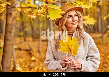 lady enjoy being in autumn forest, nature. beautiful caucasian blonde woman in hat spend time alone, walk at sunny autumn day, yellow trees and leaves around her Stock Photo