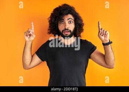 surprised arab guy in black casual t-shirt pointing finger at up, he saw something interesting, ask someone to look there. isolated orange background Stock Photo