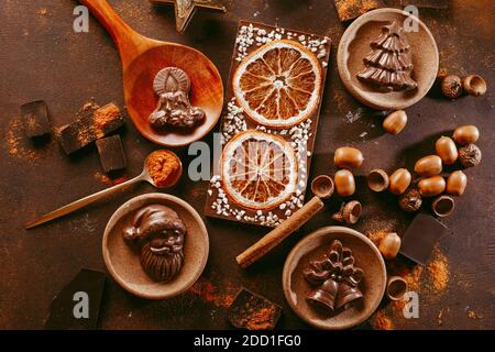 Christmas chocolate figures Santa Claus, Christmas tree, bells, candle are on the dark table next to a chocolate bar with orange and spices Stock Photo