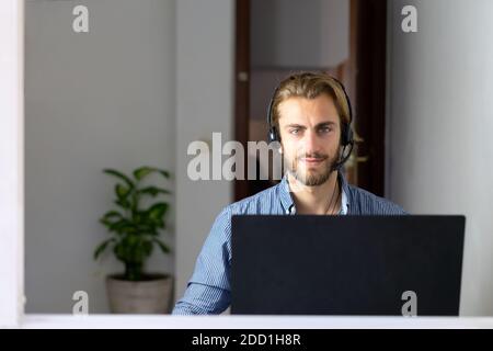 Blond male working from home with headphones using wireless internet on laptop, man call center agent or telemarketer Stock Photo