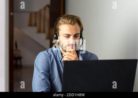Blond male working from home with headphones using wireless internet on laptop, man call center agent or telemarketer Stock Photo