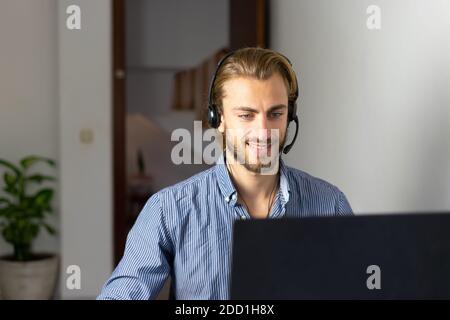 Blond male working from home with headphones using wireless internet on laptop, man call center agent or telemarketer Stock Photo