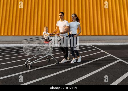 Happy family, husband and wife with child sitting in shopping trolley making purchases against yellow wall of shopping mall Stock Photo