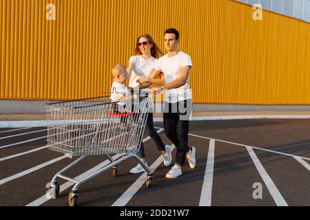 Happy family, man and woman having fun while rolling their baby in shopping trolley, outdoors, shopping concept Stock Photo