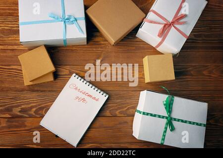 Wooden table with gift boxes and notepad with list of Christmas gifts. Space for writing. Concept of New Year. Boxes are tied with colored ribbons. Open spiral notebook. Winter holiday. Stock Photo