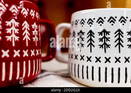 Closeup image of two (red and white) porcelain coffee mugs placed on top of table cloth. Both mugs have pine Christmas tree figures as repeating patte Stock Photo