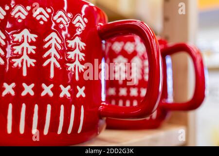 Two red shiny porcelain coffee mugs with handles placed side by side on a shelf. They have white marks and textures in shapes of pine Christmas trees. Stock Photo