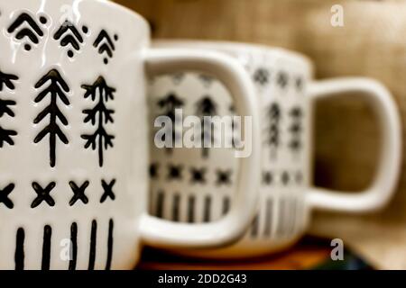 Two white porcelain coffee mugs with handles placed side by side on a shelf. They have black marks and textures in shapes of pine Christmas trees. A w Stock Photo