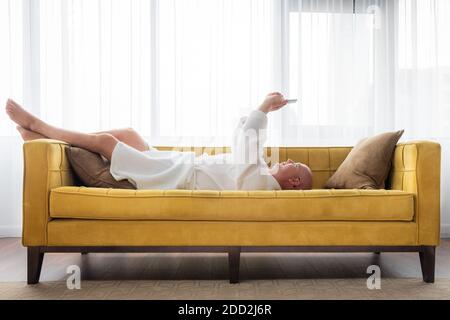 Man in white bathrobe is lying on yellow sofa and relaxing with mobilephone at home. Man chatting on mobile phone with friends. Concept stay at home. Stock Photo