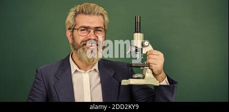 Facilitate academic progress. School teacher looking microscope. Scientific research. Fascinating research. Teacher with microscope. Man hipster classroom chalkboard background. Biological research. Stock Photo