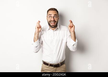 Excited and hopeful businessman making a wish, cross fingers and waiting, standing over white background Stock Photo