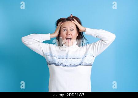 a surprised, screaming young Asian woman holds her hands to her head with her fingers in her hair, horror and fear isolated on a blue background Stock Photo