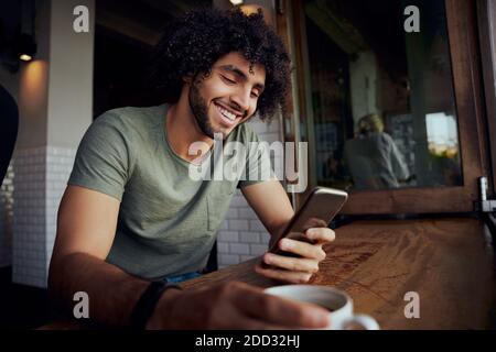 Handsome smiling young man sitting in cafe holding coffee mug while using smartphone Stock Photo