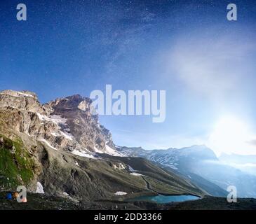 Breathtaking panoramic view of majestic mountains and blue lake under night sky with stars. Magnificent mountain landscape with starlight, rocky hills and camp tents. Concept of travelling in Alps Stock Photo