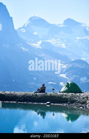 Vertical shot man tourist enjoying scenery, sitting in chair near tent and lake with fresh clear water. Rocky alpine mountains on background. Stock Photo