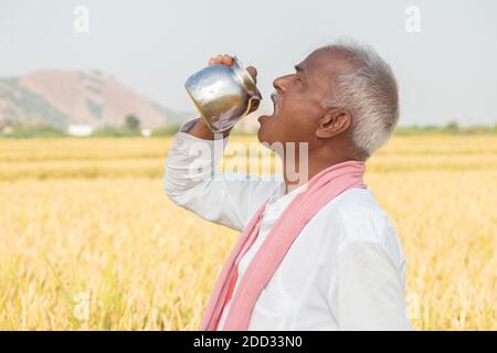 Thirsty Indian farmer drinking water from steel tumbler or Chambu on hot sunny day at agriculture field Stock Photo