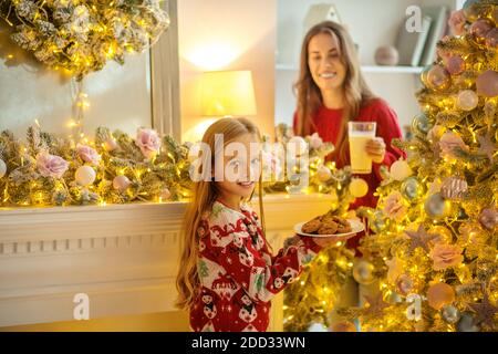 Girl holding a plate with fresh homemade pastries, her mom smiling Stock Photo
