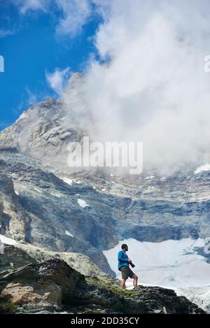 Side view of tourist in shorts standing on rocky hill and holding professional camera. Man hiker enjoying the view of beautiful snowy mountains. Concept of travelling, hiking and mountaineering. Stock Photo