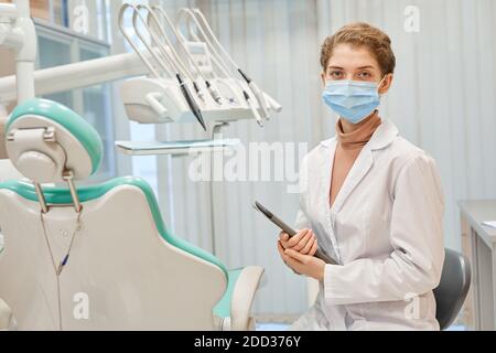Portrait of young dentist in mask and in lab coat holding digital tablet looking at camera while standing at office Stock Photo
