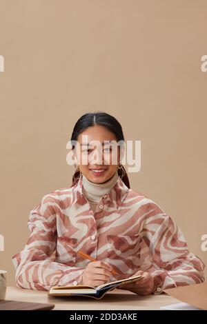Portrait of young Asian woman smiling at camera while sitting at the table and writing in note pad Stock Photo