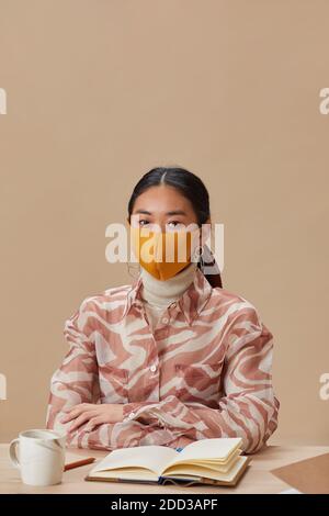 Portrait of Asian woman in protective mask looking at camera while sitting at the table and studying Stock Photo