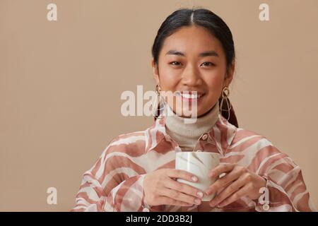 Portrait of Asian young woman smiling at camera holding mug with tea isolated on beige background Stock Photo