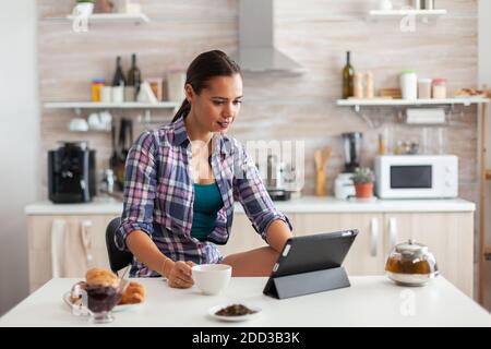 Cheerful woman smiling and reading news on tablet while drinking green tea. Working from home using device with internet technology, typing, on gadget during breakfast. Stock Photo