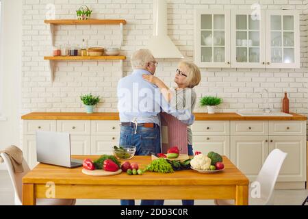 Happy elderly couple hugging or dancing in kitchen during cooking fresh healthy vegan meal Stock Photo
