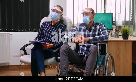 Businessman in wheelchair and woman partner with face masks having videomeeting in business office during global pandemic. Disabled freelancer working in financial company respecting social distance. Stock Photo
