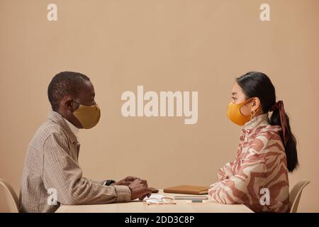 Side view of two colleagues in protective masks sitting at the table and looking at each other during meeting Stock Photo