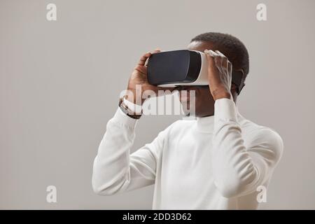 African young man in virtual glasses playing in virtual reality game against the grey background Stock Photo