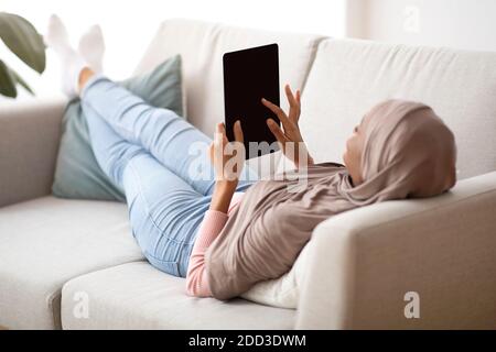 African American woman in hijab with tablet computer lying on sofa at home, mockup for design on screen Stock Photo