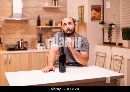 Depressed young man with alcohol addiction having wine degustation in kitchen sitting at table. Unhappy person disease and anxiety feeling exhausted with having alcoholism problems. Stock Photo