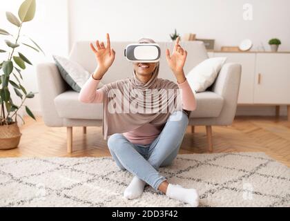Weekend pastimes. Young Muslim woman in hijab interacting with virtual reality, wearing VR headset at home Stock Photo