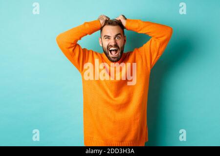 Frustrated man shouting, pulling out hair and screaming angry, losing temper and looking mad, standing over light blue background Stock Photo