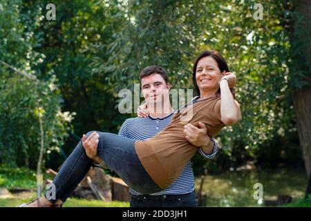 Portrait of down syndrome adult man carrying mother in arms outdoors in garden. Stock Photo