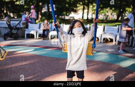 Small girl with face mask on swing outdoors in town, coronavirus concept. Stock Photo