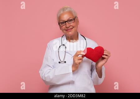 Happy smiling senior female doctor holding red heart isolated on pink background. Healthy lifestyle concept. Stock Photo