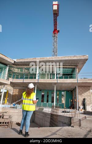 Architect supervising construction site, young female professional wearing a hardhat and neon safety jacket while holding an electronic device Stock Photo