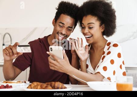 Cheerful young couple having tasty breakfast while sitting at the kitchen table, using mobile phone for shopping online with credit card Stock Photo