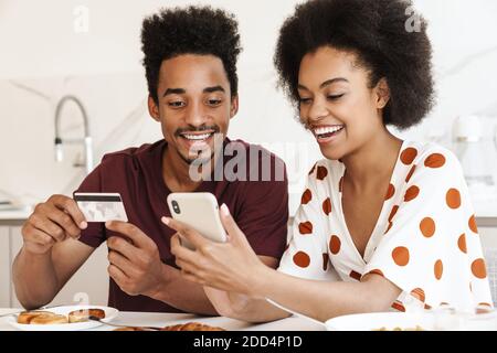 Cheerful young couple having tasty breakfast while sitting at the kitchen table, using mobile phone for shopping online with credit card Stock Photo