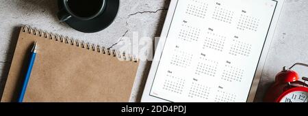 a tablet with an open calendar for 2021 year, a red alarm clock, a cup of coffee, and a craft paper notebook on a gray concrete background. banner Stock Photo