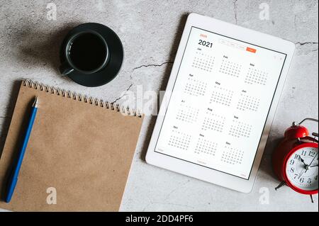 a tablet with an open calendar for 2021 year, a red alarm clock, a cup of coffee, and a craft paper notebook on a gray concrete background Stock Photo