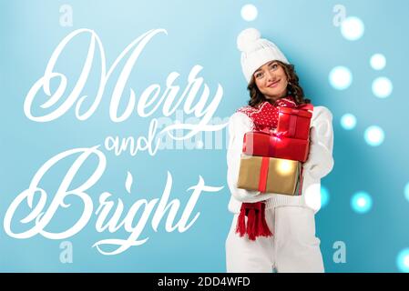 young joyful woman in winter outfit holding wrapped presents near merry and bright lettering on blue Stock Photo