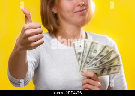 Finance, loans and banking. Close up of a young blonde holds a wad of dollars in her hands and gives a thumbs-up, smiling. Yellow background. Stock Photo