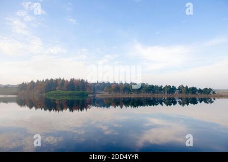 Bold mirrored landscape image of an autumnal forest. Trees form a line on the horizon, which is then reflected in the still water of a reservoir Stock Photo