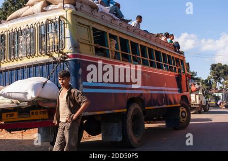 An attendant of a city bus waits for passengers in the small village of Umsning, Meghalaya, India. Passengers are seen sitting on the top of the bus. Stock Photo